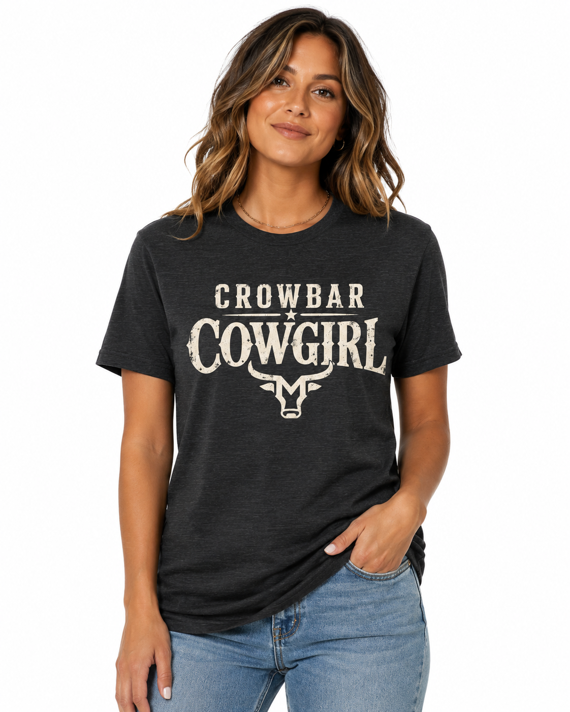 A woman wearing a dark charcoal heather t-shirt with a distressed cream graphic that reads 'CROWBAR COWGIRL' and features a steer head illustration, paired with blue jeans against a plain white background.