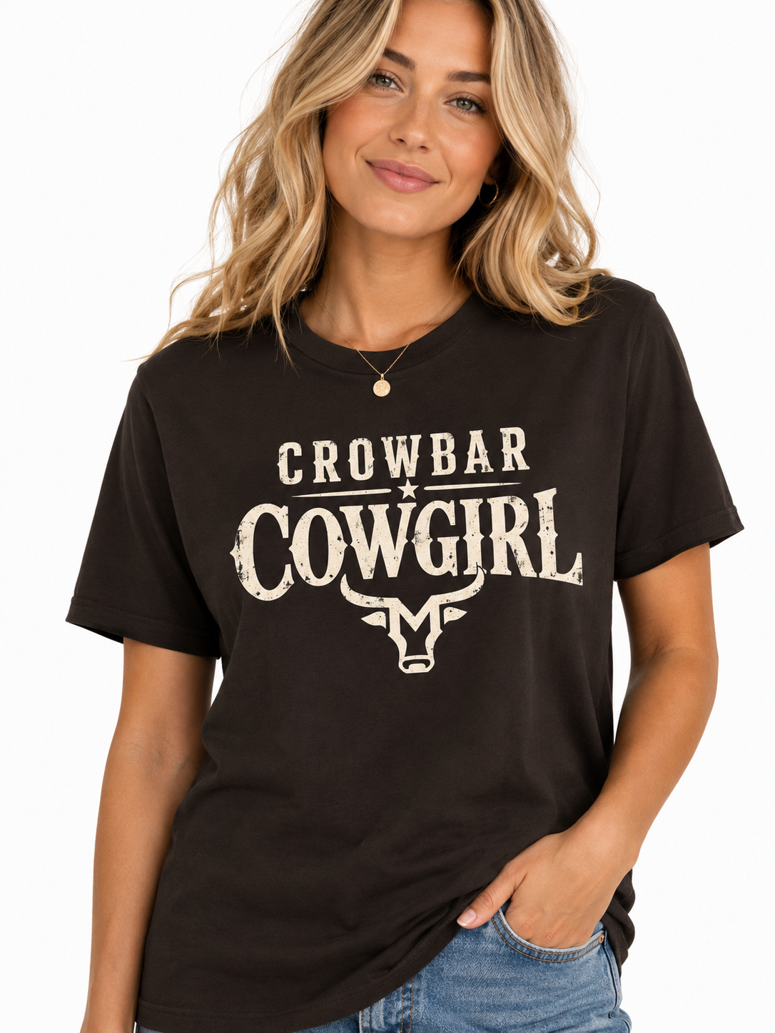 A woman wearing a brown short-sleeve t-shirt with a distressed white graphic that reads 'CROWBAR COWGIRL' and a bull skull illustration, paired with blue jeans against a plain white background.