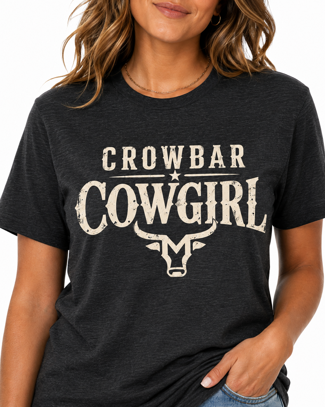 A woman wearing a dark charcoal heather t-shirt with a distressed cream graphic that reads 'CROWBAR COWGIRL' above a steer head illustration.